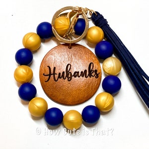 Peut inclure: Un porte-cl&eacute;s en perles dor&eacute;es et bleues avec un charme rond en bois grav&eacute; du nom "Hubanks". Le porte-cl&eacute;s a un anneau de cl&eacute; en or et un pompon bleu.