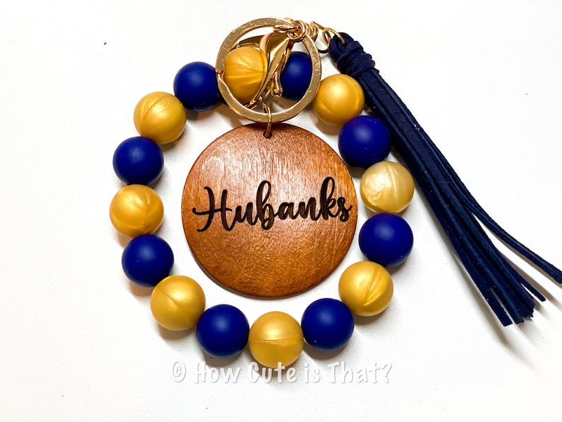 Peut inclure: Un porte-cl&eacute;s en perles dor&eacute;es et bleues avec un charme rond en bois grav&eacute; du nom "Hubanks". Le porte-cl&eacute;s a un anneau de cl&eacute; en or et un pompon bleu.