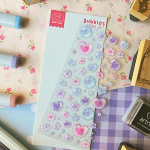 May include: A clear sticker sheet titled Bubbles Sticker Sheet by dear emzy featuring various small, translucent, iridescent stickers in shapes of circles, hearts, and stars with soft pink, blue, and purple watercolour-style gradients.