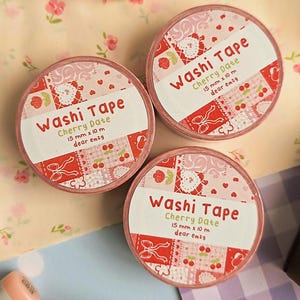 Könnte beinhalten: Drei Rollen Washi Tape mit der Aufschrift Cherry Date, jeweils 15 mm x 10 m groß, von dear emzy. Das Klebeband zeigt ein rotes und rosafarbenes Patchwork-Muster mit Herzen, Kirschen und Schleifen. Die Rollen liegen auf einer floralen Oberfläche.