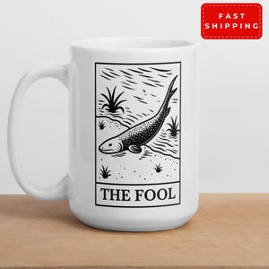 May include: White ceramic mug with a black and white tarot card design. The card depicts a fish and reads "THE FOOL". The mug has a curved handle and is on a wooden surface. The mug has a "FAST SHIPPING" sticker.