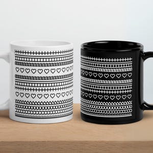 May include: Two ceramic mugs, one white and one black, with a repeating geometric pattern in white. The pattern includes hearts, lines, and other shapes. The mugs are on a wooden surface.
