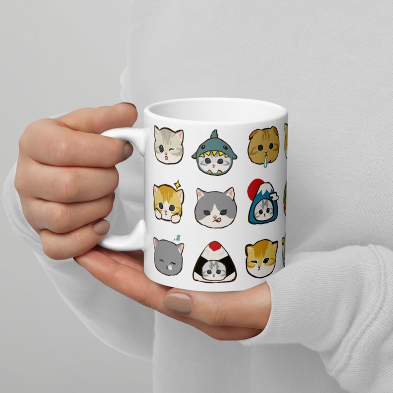Puede incluir: Taza de cer&aacute;mica blanca con un dise&ntilde;o colorido de varias caras de gato. Las caras de gato est&aacute;n en diferentes poses y expresiones, incluyendo un tibur&oacute;n, un rollo de sushi y una monta&ntilde;a.