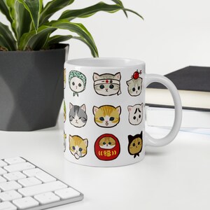 Puede incluir: Taza de cer&aacute;mica blanca con un patr&oacute;n de lindos gatos de dibujos animados en varias poses y atuendos. Los gatos llevan sombreros, bufandas y otros accesorios. La taza tambi&eacute;n presenta un amuleto de la suerte japon&eacute;s rojo y dorado con la palabra "福" (que significa "buena fortuna") escrita en &eacute;l.