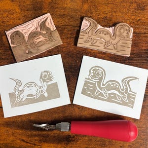 May include: Two rectangular linoleum carving blocks with carved designs of a creature, two corresponding white paper prints showing the creature in brown ink, and a carving tool with a red handle and a metal blade tip.