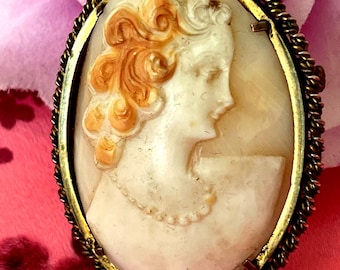 May include: Oval cameo brooch featuring a profile of a woman with detailed hair and a pearl necklace. The cameo is set in a gold-toned frame with a twisted rope border. The woman's hair is a light orange color.