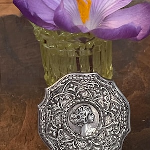 May include: An octagonal silver compact with an embossed floral design and a central cameo of a woman's profile. A purple crocus flower with an orange center sits in a small, yellow-green glass vase.