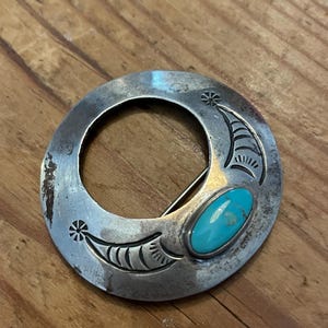 May include: A vintage silver-toned brooch with a circular design. The brooch features a turquoise oval stone and etched details. The metal has a worn appearance, suggesting age. The brooch is set on a wooden surface.