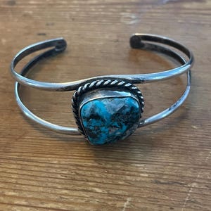 May include: Silver cuff bracelet featuring a large, square turquoise stone. The stone is set in a decorative bezel with a twisted rope border. The bracelet has a double-band design and is displayed on a wooden surface.