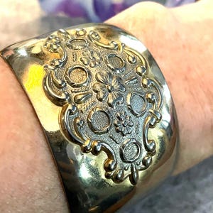 May include: Wide, silver-toned cuff bracelet with an ornate, raised floral and scroll design. The bracelet has a polished finish and a curved shape, suitable for the wrist. A statement piece of jewelry.