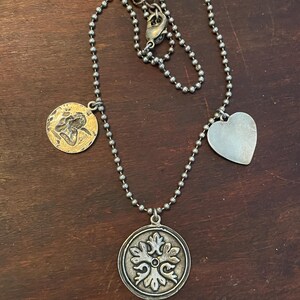 May include: Silver-toned necklace with a ball chain and multiple charms. The charms include a heart, a round pendant with a floral design, and a gold-toned coin with a figure. The necklace is displayed on a dark brown wooden surface.