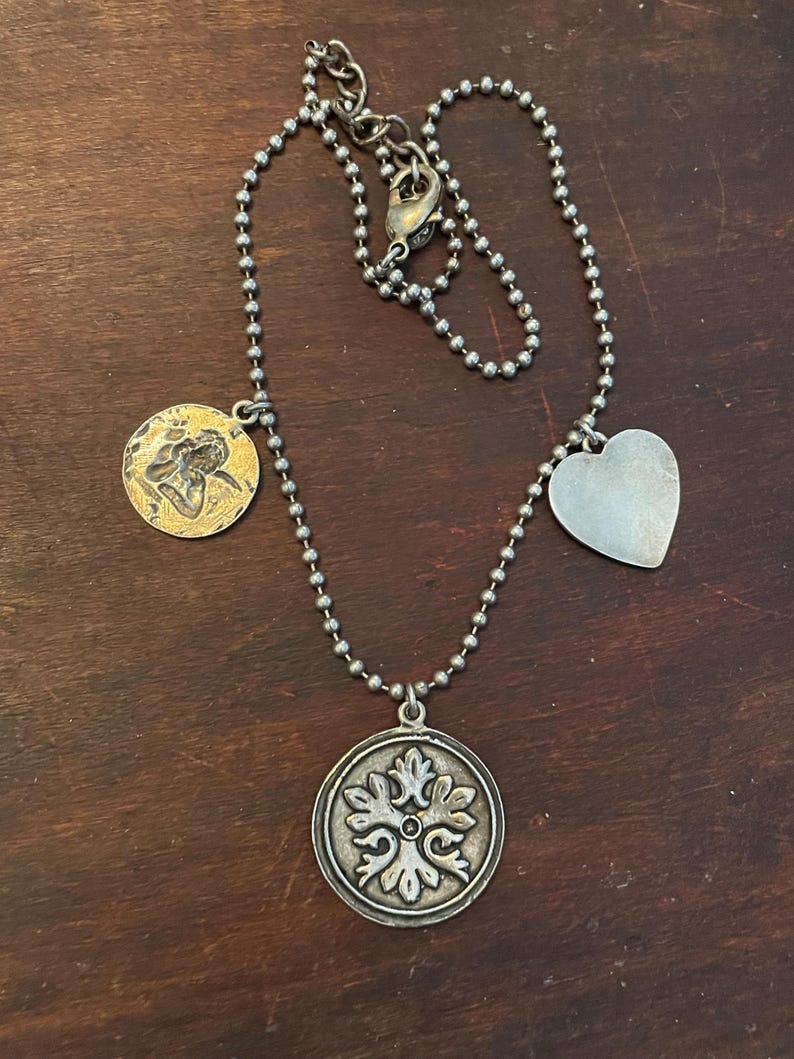 May include: Silver-toned necklace with a ball chain and multiple charms. The charms include a heart, a round pendant with a floral design, and a gold-toned coin with a figure. The necklace is displayed on a dark brown wooden surface.