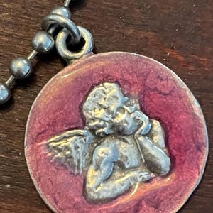May include: A silver-toned pendant featuring a cherub design within a circular, maroon-colored enamel setting. The pendant is attached to a ball chain necklace, creating a vintage aesthetic.
