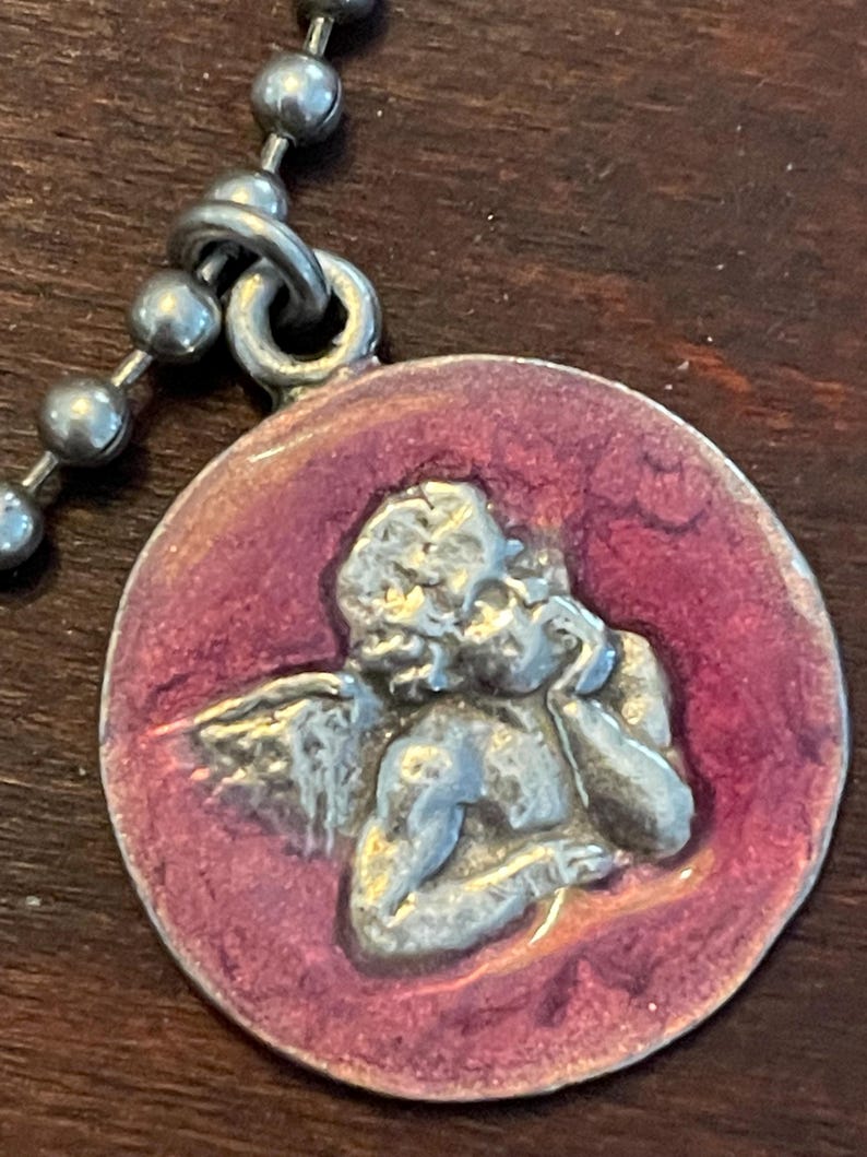 May include: A silver-toned pendant featuring a cherub design within a circular, maroon-colored enamel setting. The pendant is attached to a ball chain necklace, creating a vintage aesthetic.