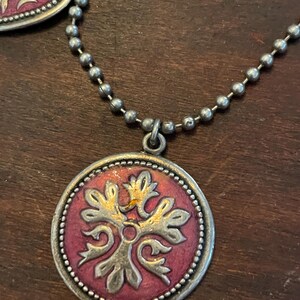 May include: A silver-toned necklace with a round pendant featuring a red center and a gold and silver floral design. The necklace has a ball chain and a matching heart-shaped pendant is partially visible.