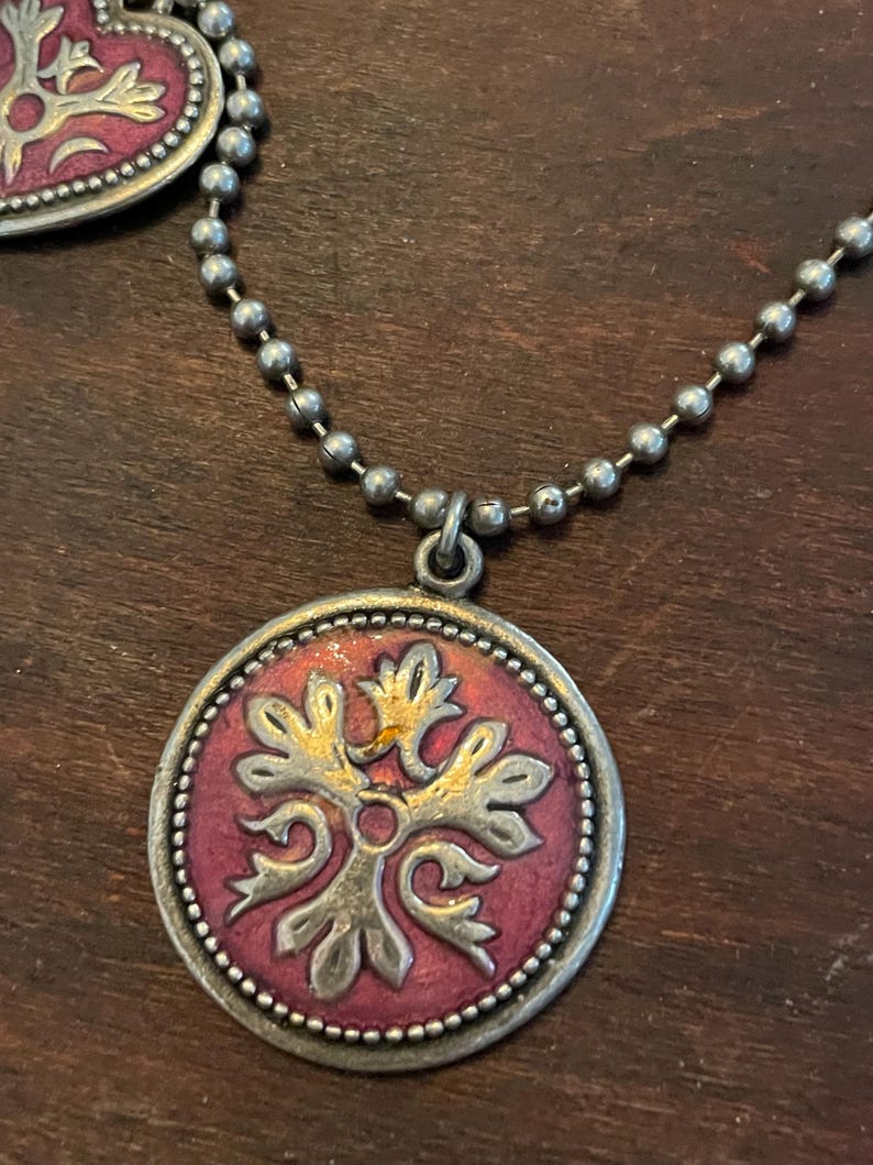 May include: A silver-toned necklace with a round pendant featuring a red center and a gold and silver floral design. The necklace has a ball chain and a matching heart-shaped pendant is partially visible.