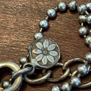 May include: Close-up of a silver necklace with a daisy-shaped clasp. The necklace features a ball chain design and a lobster clasp. The clasp has a daisy flower design in the center.