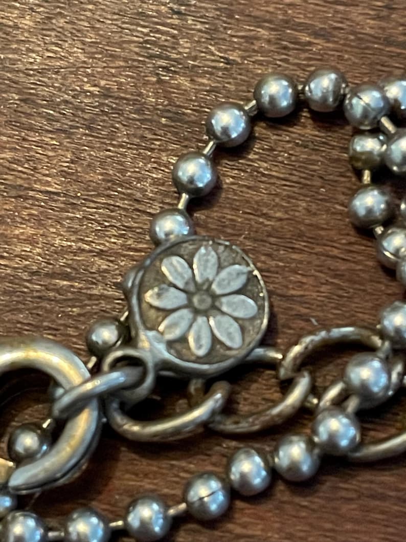 May include: Close-up of a silver necklace with a daisy-shaped clasp. The necklace features a ball chain design and a lobster clasp. The clasp has a daisy flower design in the center.
