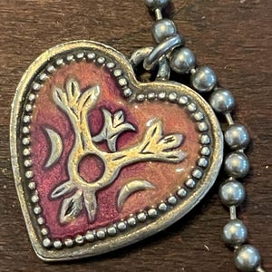May include: A heart-shaped pendant with a silver-colored border and a ball chain. The pendant features a gold-colored floral design over a red and pink background. The border is dotted with small silver beads.