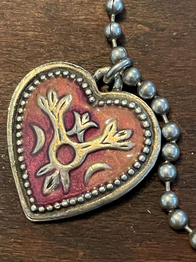 May include: A heart-shaped pendant with a silver-colored border and a ball chain. The pendant features a gold-colored floral design over a red and pink background. The border is dotted with small silver beads.