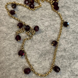 Pode incluir: Um delicado colar dourado com uma série de contas facetadas de cor vermelho escuro. As contas são intercaladas ao longo da corrente, criando um efeito gracioso e drapeado. O colar tem um design clássico, adequado para várias ocasiões.