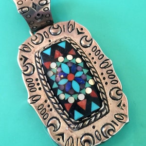 May include: A silver pendant featuring a rectangular inlay of turquoise, coral, and other stones set in black. The pendant is surrounded by a silver border with a repeating crescent moon design.