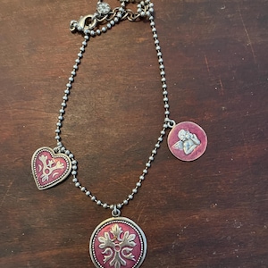 May include: A silver-toned necklace with three pendants. The pendants include a heart, a round pendant with a floral design, and a round pendant with an angel. The pendants have a red background with silver-toned designs.