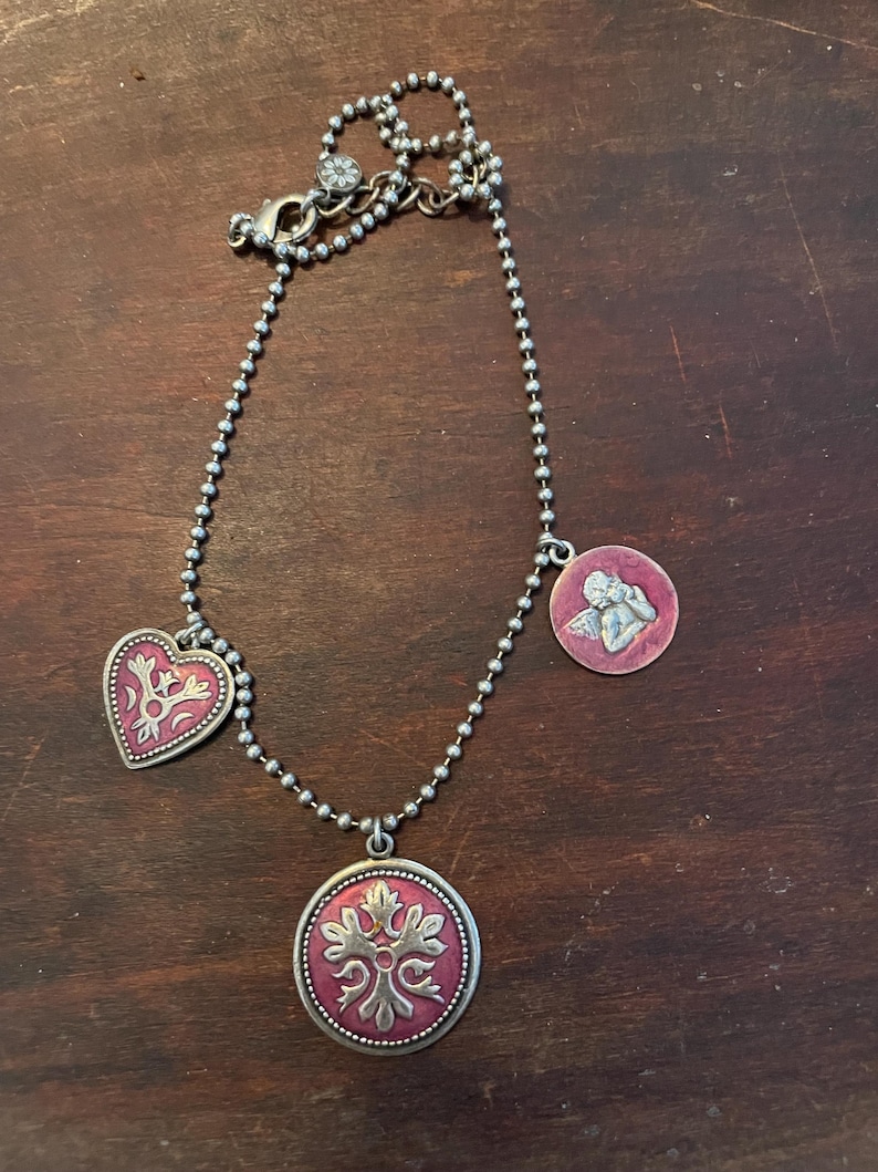May include: A silver-toned necklace with three pendants. The pendants include a heart, a round pendant with a floral design, and a round pendant with an angel. The pendants have a red background with silver-toned designs.