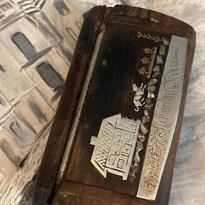 Può includere: Una scatola rettangolare in legno con finitura scura, con elementi decorativi in metallo e intarsi. La scatola ha una placca metallica color argento con la scritta "LA BRETINE" e un disegno in metallo a forma di casa. La scatola è probabilmente vintage.