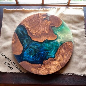 May include: Round wooden serving board with an ocean-inspired resin design. The board features natural wood grain and a vibrant mix of blue, teal, and white resin, creating a coastal aesthetic. The text "Ocean-Milano Effect" is visible on the left side.