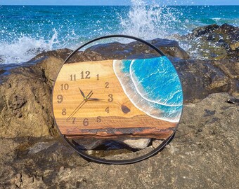 May include: A round wooden clock with a blue and white resin ocean wave design. The clock face has black numbers and hands. The clock is sitting on a rocky surface.