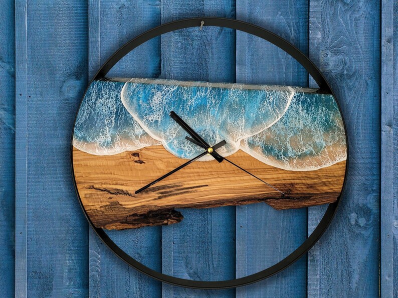 Pu&ograve; includere: Un orologio rotondo in legno con cornice nera. Il quadrante dell'orologio &egrave; in legno marrone chiaro con un design a onda in resina blu e bianca. Le lancette dell'orologio sono nere.