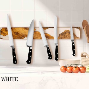 May include: Five silver kitchen knives with black handles are mounted on a wooden magnetic strip with a white background. The strip has a light brown wood grain pattern and a white epoxy resin inlay. The knives are arranged in a row, with the largest knife on the left and the smallest knife on the right. The knives are all sharp and have a polished finish. The word "WHITE" is printed in black letters on the bottom left corner of the image.