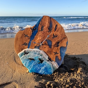 May include: A circular wall clock crafted from a natural wood slab with blue resin accents resembling water. The clock face features embedded black bird silhouettes and a textured, white-capped blue resin wave design at the bottom.