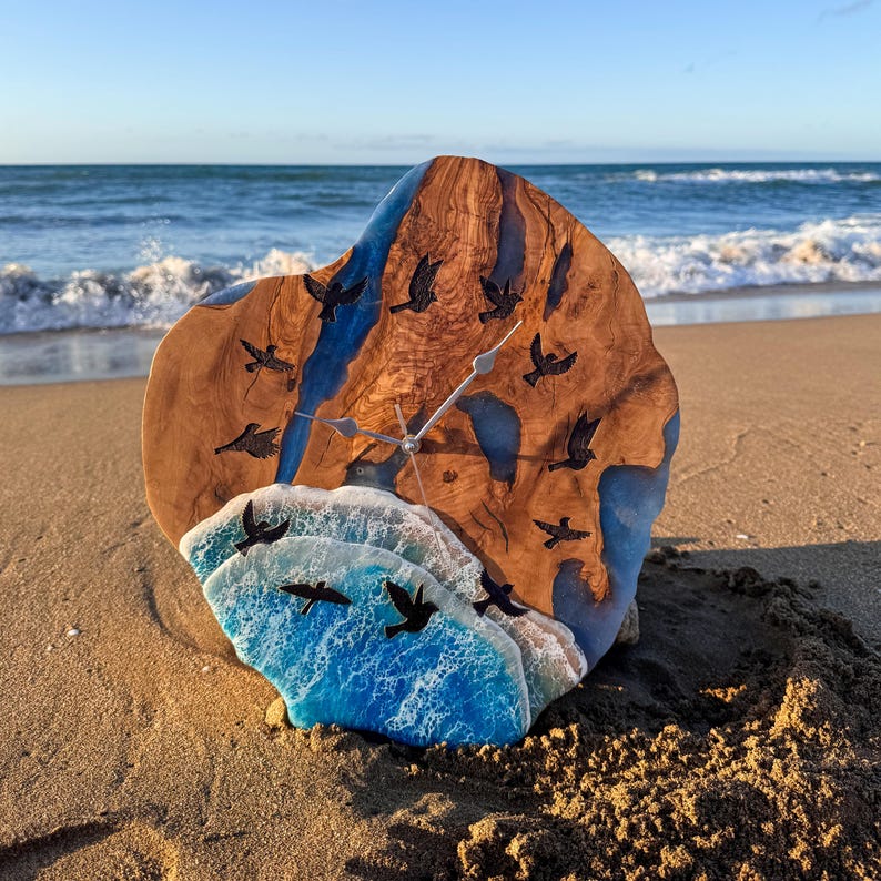 May include: A circular wall clock crafted from a natural wood slab with blue resin accents resembling water. The clock face features embedded black bird silhouettes and a textured, white-capped blue resin wave design at the bottom.