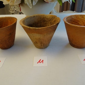 Könnte beinhalten: Drei Terrakotta-Töpfe in verschiedenen Größen, beschriftet mit "S", "M" und "L" auf weißen Papierquadraten. Die Töpfe sind unglasiert und haben ein rustikales, erdiges Aussehen.