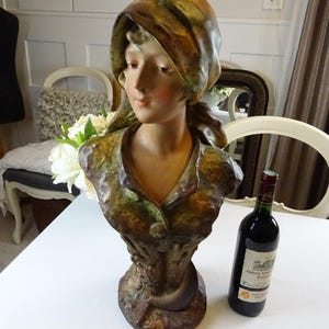 May include: A ceramic bust of a woman with a brown and green patterned dress and a brown headscarf. The bust is on a table with a bottle of wine.