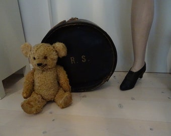 May include: A brown teddy bear with a black nose and button eyes sits next to a round, black, leather suitcase with the initials "R.S." on the side.