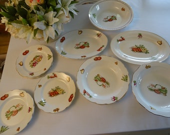 May include: A set of nine white ceramic plates with a gold rim and a vegetable design. The plates are in various sizes, including a large oval platter, a smaller oval platter, and several round plates.