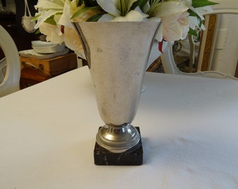 May include: A silver trophy cup with a black marble base. The cup is shaped like a vase and has a round base.