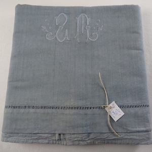 Könnte beinhalten: Eine hellblaue Tischdecke aus Leinen mit einem weißen gestickten Monogramm "A M" und einem kleinen handgeschriebenen Etikett, das mit einer Schnur befestigt ist.