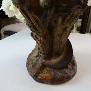 May include: A brown ceramic vase with a sculpted design of wheat stalks and a crescent moon. The vase has a textured surface and a rustic appearance.