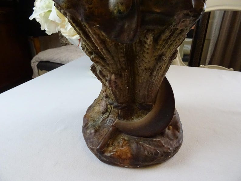 May include: A brown ceramic vase with a sculpted design of wheat stalks and a crescent moon. The vase has a textured surface and a rustic appearance.