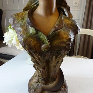 May include: A ceramic bust sculpture of a woman with a green and brown patterned dress. The bust is on a base with a crescent moon design.