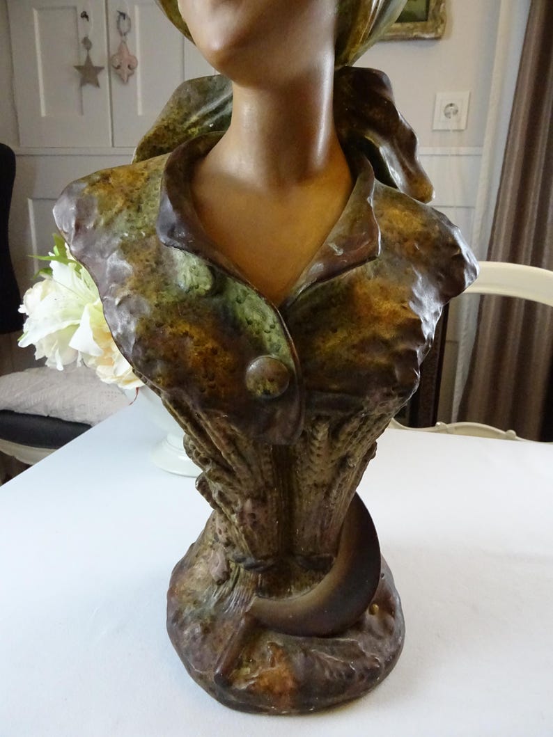 May include: A ceramic bust sculpture of a woman with a green and brown patterned dress. The bust is on a base with a crescent moon design.