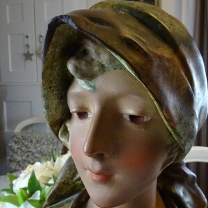 May include: A ceramic bust of a woman with a brown and green draped fabric covering her head. The bust is painted with realistic skin tones and features.