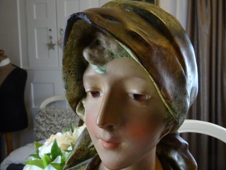 May include: A ceramic bust of a woman with a brown and green draped fabric covering her head. The bust is painted with realistic skin tones and features.