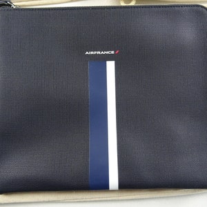Pode incluir: Uma bolsa preta com zíper com uma faixa branca e azul no centro. A bolsa tem o logotipo da Air France em letras vermelhas na frente.
