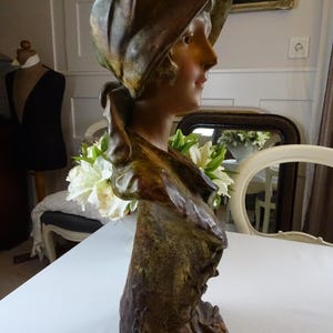 May include: A bronze bust sculpture of a woman wearing a draped hat. The sculpture is painted in shades of brown and green, and the woman's face is delicately sculpted.