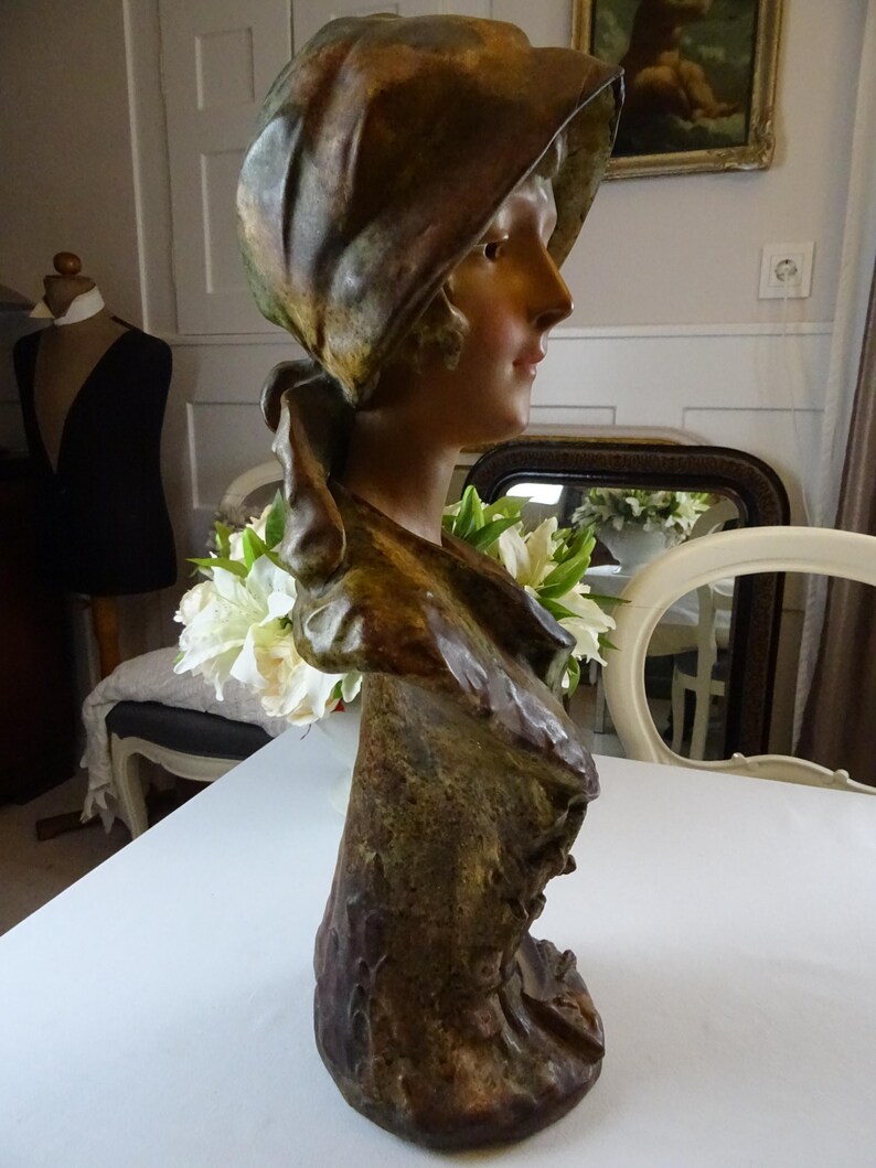 May include: A bronze bust sculpture of a woman wearing a draped hat. The sculpture is painted in shades of brown and green, and the woman's face is delicately sculpted.